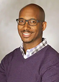 Elon is a bald man with light facial hair and glasses. He is wearing a purple sweather and patterned shirt.