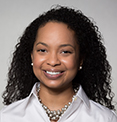 Shanele has medium-length curly black hair. She is wearing a white shirt and a beaded necklace.