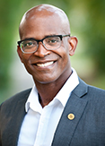 Wayne is a bald man wearing black-rimmed glasses and is smiling.