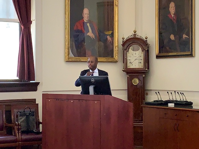 Wayne Frederick stands at the lectern in the Waterhouse Room. There is a grandfather clock and two large paintings behind him.