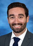 Michael Harding has short black hair and facial hair. He is wearing a black tie, white shirt, and black blazer.
