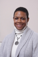 Sherri-Ann Burnett-Bowie is wearing a light gray cardigan, white turtleneck, and pendant necklace. She has short cropped black hair.