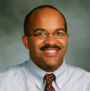 Abner Louissaint is a balm man with a moustache. He is wearing a light grey shirt and red patterned tie.
