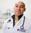 Alister Francois Martin is standing in a doctor's office. He is wearing a white doctor's coat and stethoscope.