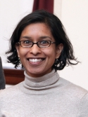 Sudha Biddinger has short black hair and glasses. She is wearing a beige turtleneck.