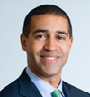 William Curry has very short dark hair. He is wearing a black blazer, blue shirt, and green tie.
