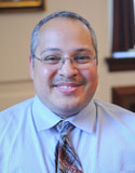 Elliot Melendez is wearing a white shirt and patterned tie. He has short gray hair and glasses.