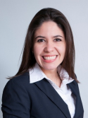 Lidia Moura is wearing a black blazer and white shirt. She has long brown hair.