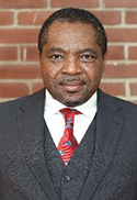 Matthew Yuyun is wearing a 3-piece suit with a red patterned tie. He is standing in front of a brick wall.