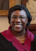Olivia Okereke is wearing a magenta blouse and black jacket. She is standing in front of a framed painting and a clock.