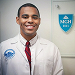 Randy Miles is wearing a white coat, white shirt, and red tie. He is standing next to an MGH sign.