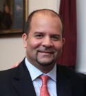 Aaron Kyle Styer is a bald man with a goatee. He is wearing a black blazer, blue shirt, and red tie.