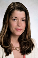 Viviany Rodrigues Taqueti has long brown and blonde hair. She is wearing a white blazer, orange blouse, and chain necklace.