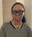 Ayobami Akenroye is standing in front of a wall. She is wearing a pearl necklace and black-and-white patterned top, as we all thick framed glasses.