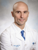 Luis Gonzalez Castro is a bald man wearing a doctor's coat, white shirt, and blue tie.