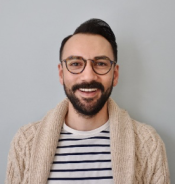 Michael Flores is wearing a beige cardigan and striped shirt. He has short brown hair, a beard and moustache, and glasses.