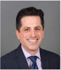 Oren Ganor has short black hair and is wearing a black blazer, blue tie, and lavender shirt.