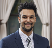 Nathaniel Harnett is standing in front of white curtains. He is wearing a blue blazer, striped white shirt, and gray striped tie.
