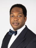 Asishana Osho is wearing a black blazer, white shirt, and black bowtie.