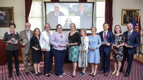  11 recipients of the 2025 Mentoring Awards pose with their awards in the Waterhouse Room. Three others are pictured virtually on a projector in the back.