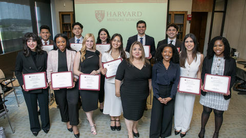 Eleven VRIP students pose with their certificates alongside two staff members. The Harvard Medical School logo is projected on a screen behind them. 