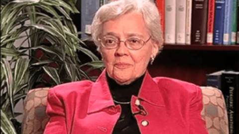 Eleanor Shore is wearing a pink jacket and black top. There is a plant and bookshelf behind her.