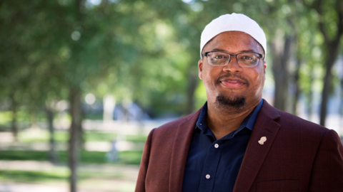 Khalil Abdur-Rashid stands outside by some trees and wears a white kufi, burgundy blazer, and dark blue button down shirt. 