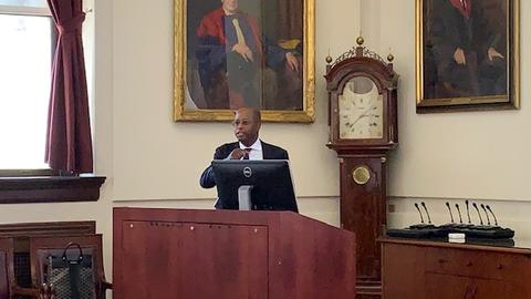 Wayne Frederick stands at the lectern in the Waterhouse Room. There is a grandfather clock and two large paintings behind him.
