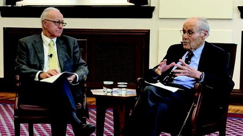 HMS Dean George Daley sits next to David Nathan. There is a table with two glasses of water between them.