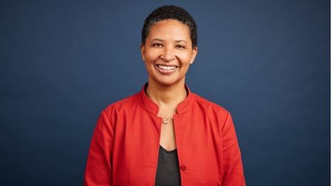 Danielle Allen stands in front of a blue background wearing a red jacket and black top. She has short black hair and is smiling.