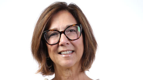 Tracy Battaglia has a brown bob and eyeglasses. She is wearing a white blouse and has her arms crossed.