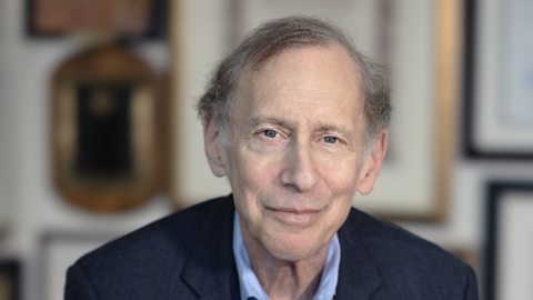 Bob Langer is wearing a dark blue blazer and light blue shirt. He is sitting in a chair with his hands clasped. There are frames on the wall behind him.