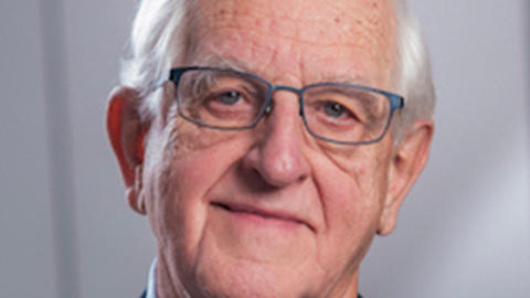 Joseph Martin has short white hair and is wearing a blue shirt with a dark blue blazer. He also has blue-framed glasses.