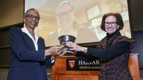 Melissa Maher is on a projector screen while two people present her the Federman award at an HMS town hall meeting.
