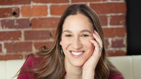 Rachel Fish is sitting on a white couch with a brick wall background. She is wearing a maroon blouse and blue jeans.