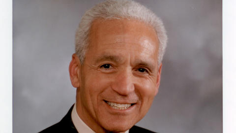 Gus White has short grey hair and is wearing a black blazer, white shirt, and brown tie.