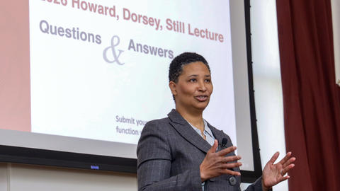Danielle Allen stands in front of a projector screen in the Waterhouse Room. Both her hands are raised in gesture as she speaks.