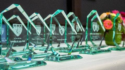 Polygonal-shaped glass awards are displayed a table with a white tablecloth. There is a vase of colorful flowers in the background.