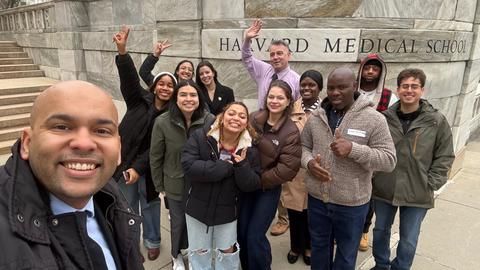 Pablo da Silva takes a selfie in front of the Harvard Medical School sign on the Quad. He is standing with a BHCC professor and 10 BHCC students, some of whom are throwing peace signs or waving.