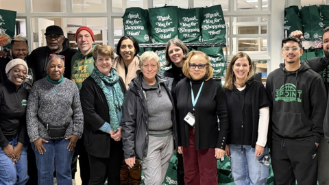 The Dimock Center and OCCE crew stands in front of green reusable bags of packaged foods. The bags say, "Thank You For Shopping Here!"