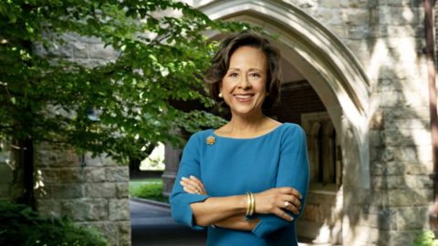 Paula Johnson is wearing a teal dress and gold accessories. She is standing outside a stone arch with greenery on either side. 