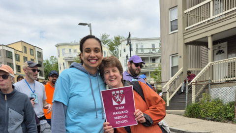 Two members of the OCCE staff stand outside during the Mother's Day Walk. One of them is holding up a red Team Harvard Medical School sign.
