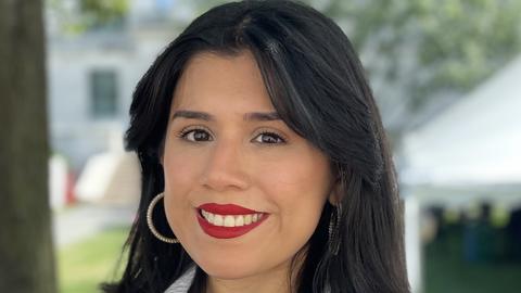 Genesis Perez-Melara is wearing a black blouse and white coat. She is standing outside in front of a tree.