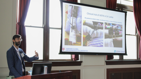 Kiran Agarwal-Harding stands behind a lectern as he gestures towards a projecter screen in the Waterhouse Room of Gordon Hall.