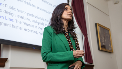 Monica Bharel is wearing a green blazer and necklace made of kukui nuts. She is standing in front of a projector screen in the Waterhouse Room and speaking.