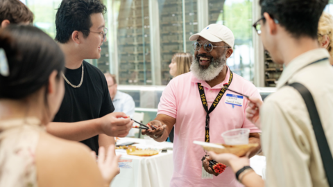 A group of people stand in a circle while a man in a pink polo in the middle passes out pencils.