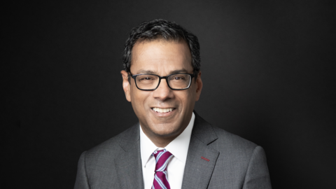 Atul Gawande is posing with his hand across his lap. He is wearing a gray suit, striped tie, and glasses.