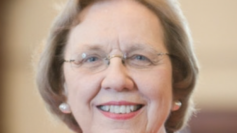 Barbara McNeil has short blonde hair and is wearing glass, pearl earrings, and a red blazer.