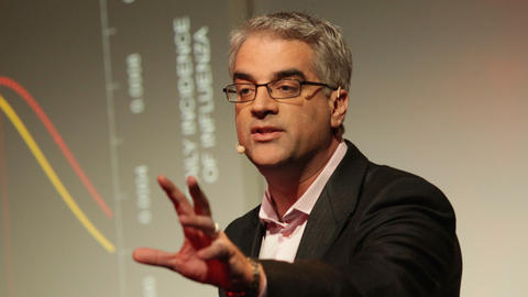 Nicholas Christakis has short gray hair and glasses. He is gesturing away from himself as he stands in front of a dark screen showing a graph.