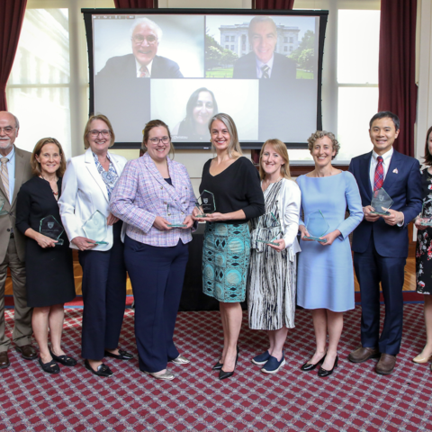  11 recipients of the 2025 Mentoring Awards pose with their awards in the Waterhouse Room. Three others are pictured virtually on a projector in the back.